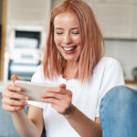 Image of delighted woman playing video game on mobile phone while sitting on sofa at home