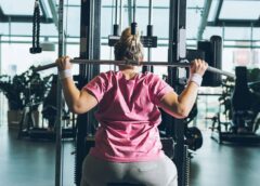 An overweight woman in a pink t-shirt flexing her muscles while using resistance training gym equipment. Exercise such as resistance training helps diet-resistant women improve their fitness.