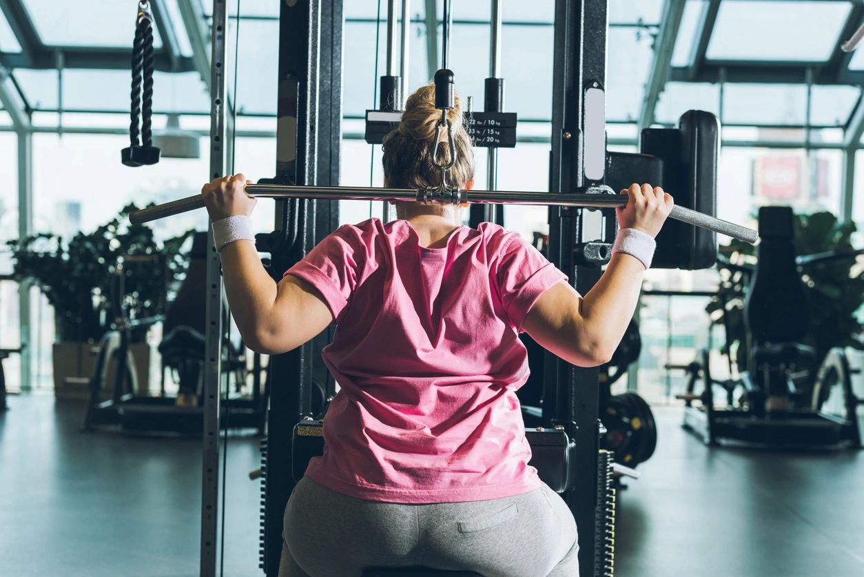 An overweight woman in a pink t-shirt flexing her muscles while using resistance training gym equipment. Exercise such as resistance training helps diet-resistant women improve their fitness.