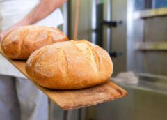 Two loaves of freshly baked bread held out on a wooden baker's paddle, with the baker and commercial oven obscured in background.