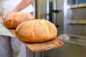 Two loaves of freshly baked bread held out on a wooden baker's paddle, with the baker and commercial oven obscured in background.