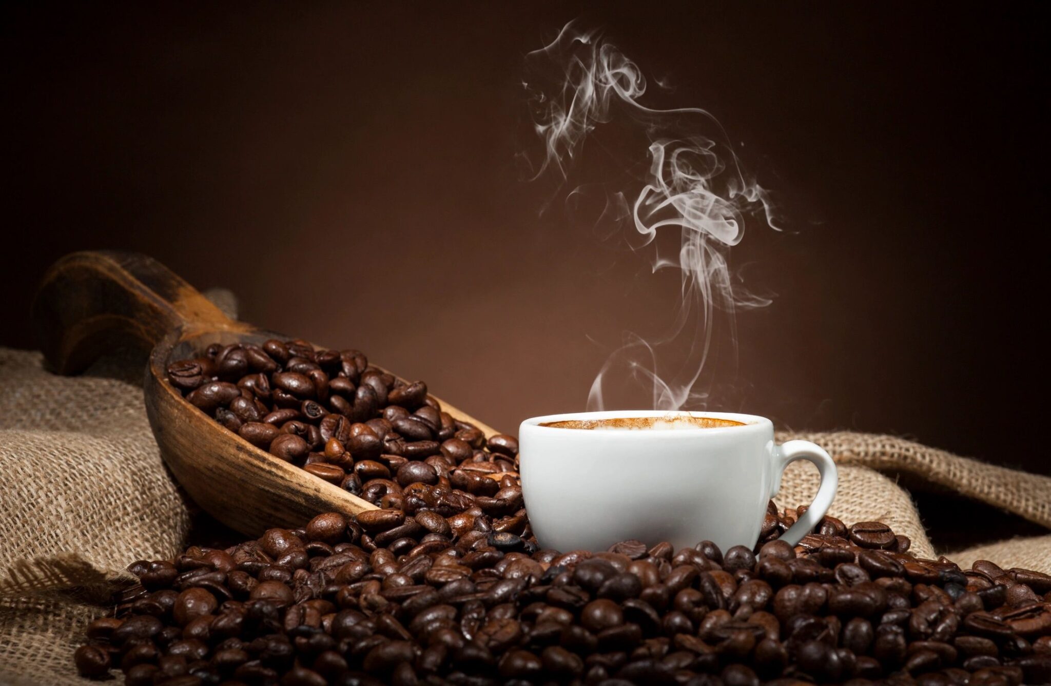 Coffee beans pour out of an open scoop onto a table next to a steaming cappuccino cup