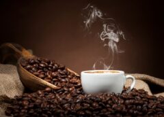 Coffee beans pour out of an open scoop onto a table next to a steaming cappuccino cup