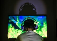 Solo videogamer with over-ear headphones sits with back to the camera in front of a lit-up television screen