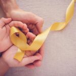Two sets of hands hold a yellow ribbon formed in a loop for cancer awareness.