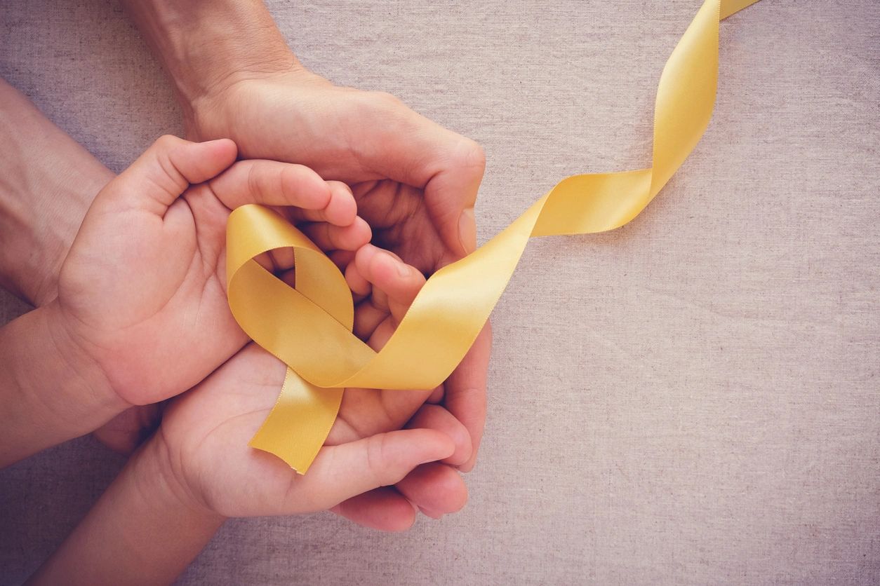 Two sets of hands hold a yellow ribbon formed in a loop for cancer awareness.