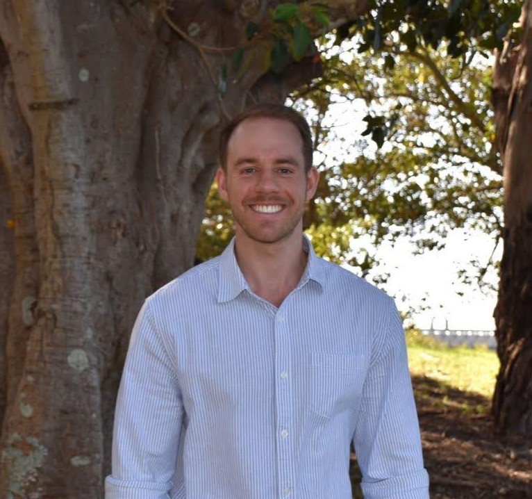 Author photo of a smiling white male wearing a white button-down shirt and standing outside in front of a large tree