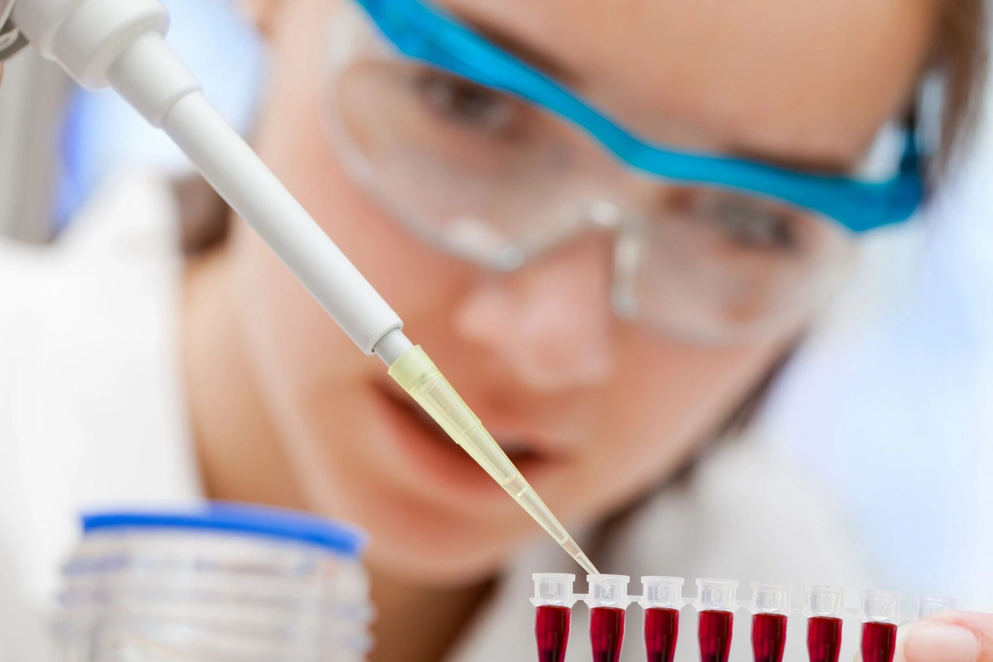 Female scientist with safety goggles holds a pipette over a row of small vials of blood